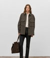 A model stands against a light gray studio background wearing an oversized brown and black houndstooth button-up shirt jacket over a cream shirt, paired with dark charcoal jeans and holding a large brown suede tote bag.