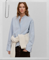 A woman modeling a light blue long-sleeve button-up shirt with a stand collar, paired with a cream ribbed knit sweater wrapped around her waist and dark brown pants, set against a light gray background.