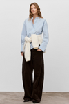 A woman stands against a plain light gray studio wall wearing a light blue button-up shirt, dark brown wide-leg corduroy pants, and a cream ribbed knit wrap tied around her waist.