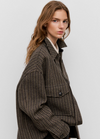 A woman wearing an oversized brown and black houndstooth patterned jacket over a white collared shirt, photographed against a light gray studio background.