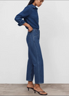 A model wearing a dark blue denim shirt and matching wide-leg cropped jeans, paired with black heeled sandals, shown from a side profile against a plain light gray wall.