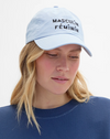 A woman wearing a light blue baseball cap with the black embroidered text 'MASCULIN FÉMININ' on the front, paired with a navy blue crewneck sweatshirt against a plain white background.